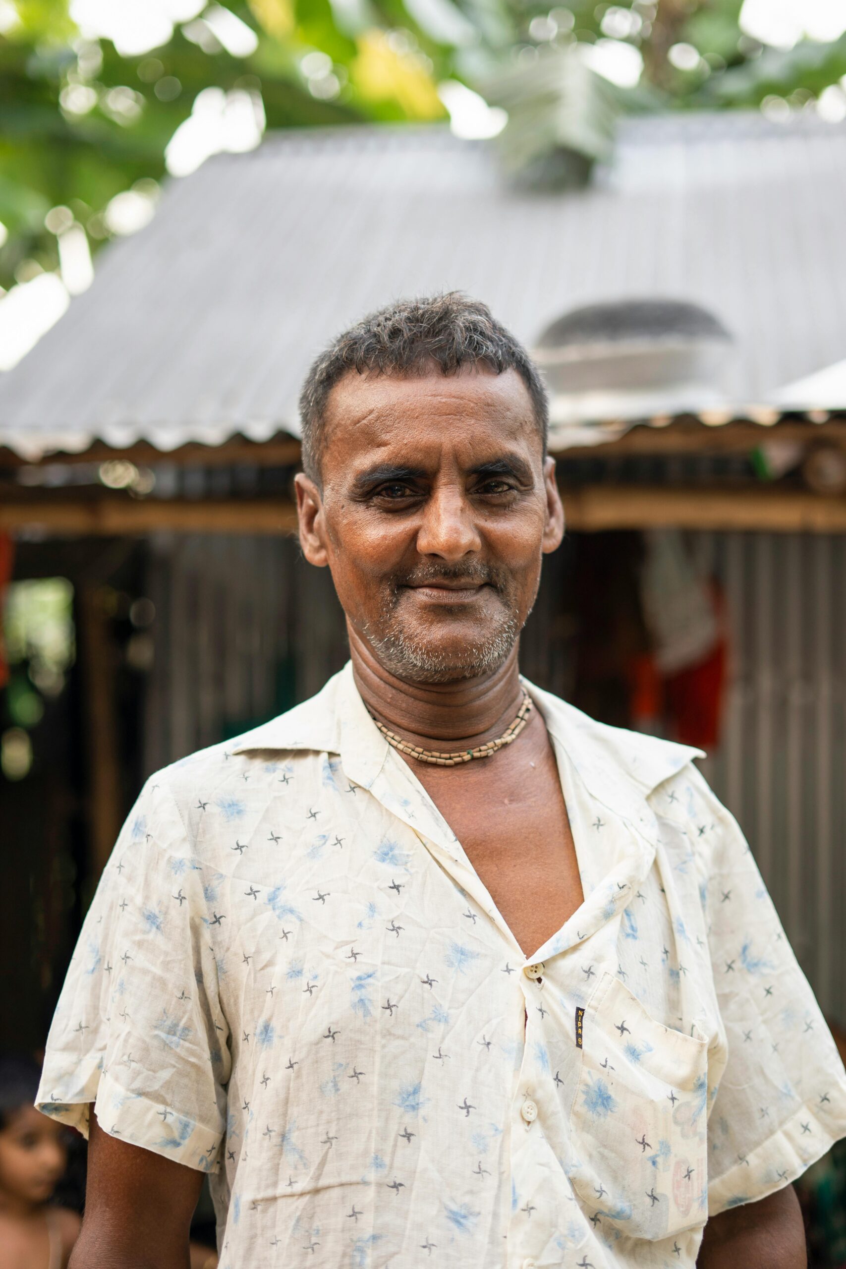 A candid portrait of a South Asian man wearing a gold necklace, captured outdoors in natural light.